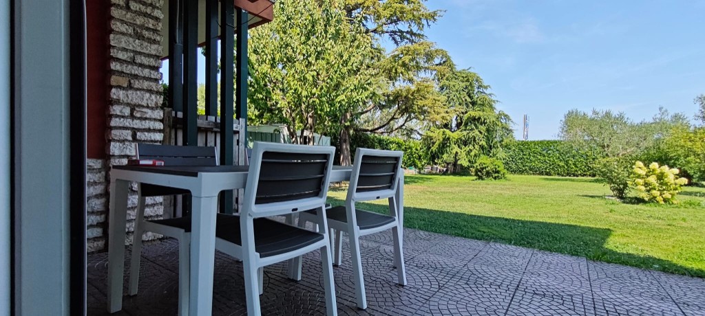 Appartamento 2.Rosso — patio con tavolo e sedie sotto il portico, affacciato sul giardino con cedro e siepe